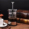 Compact stovetop moka pot coffee maker with glass top and stainless steel base.