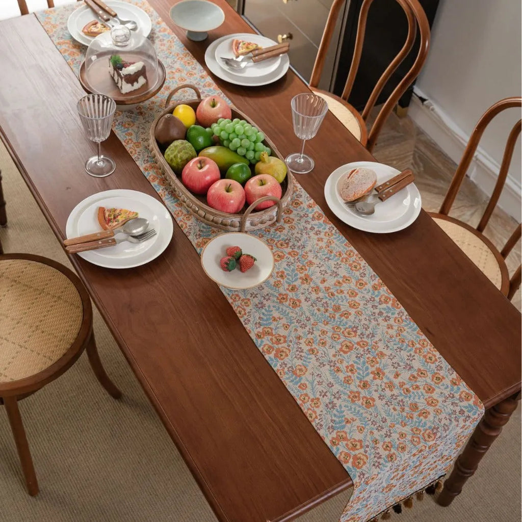 Long beige jacquard table runner with floral pattern and tassels on a wooden dining table.