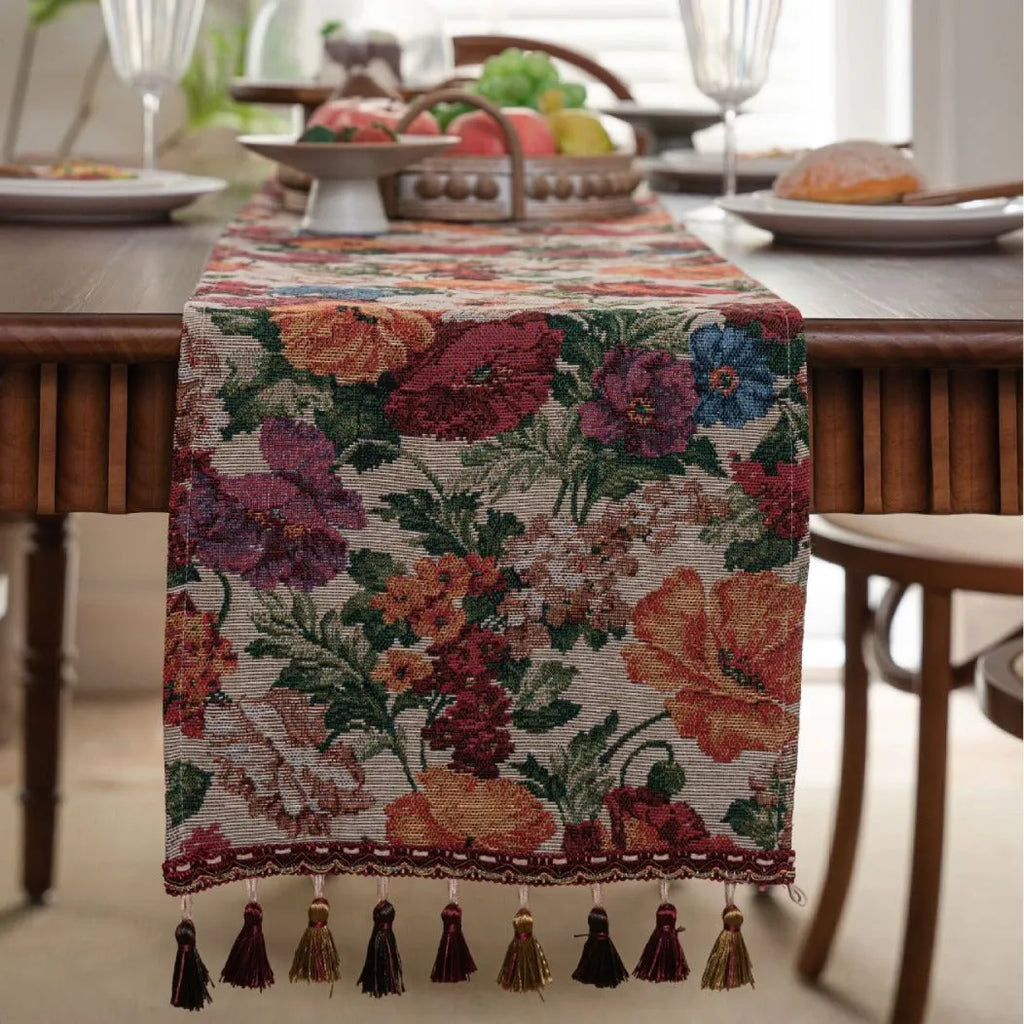 Long beige jacquard table runner with floral pattern and tassels on a wooden dining table.