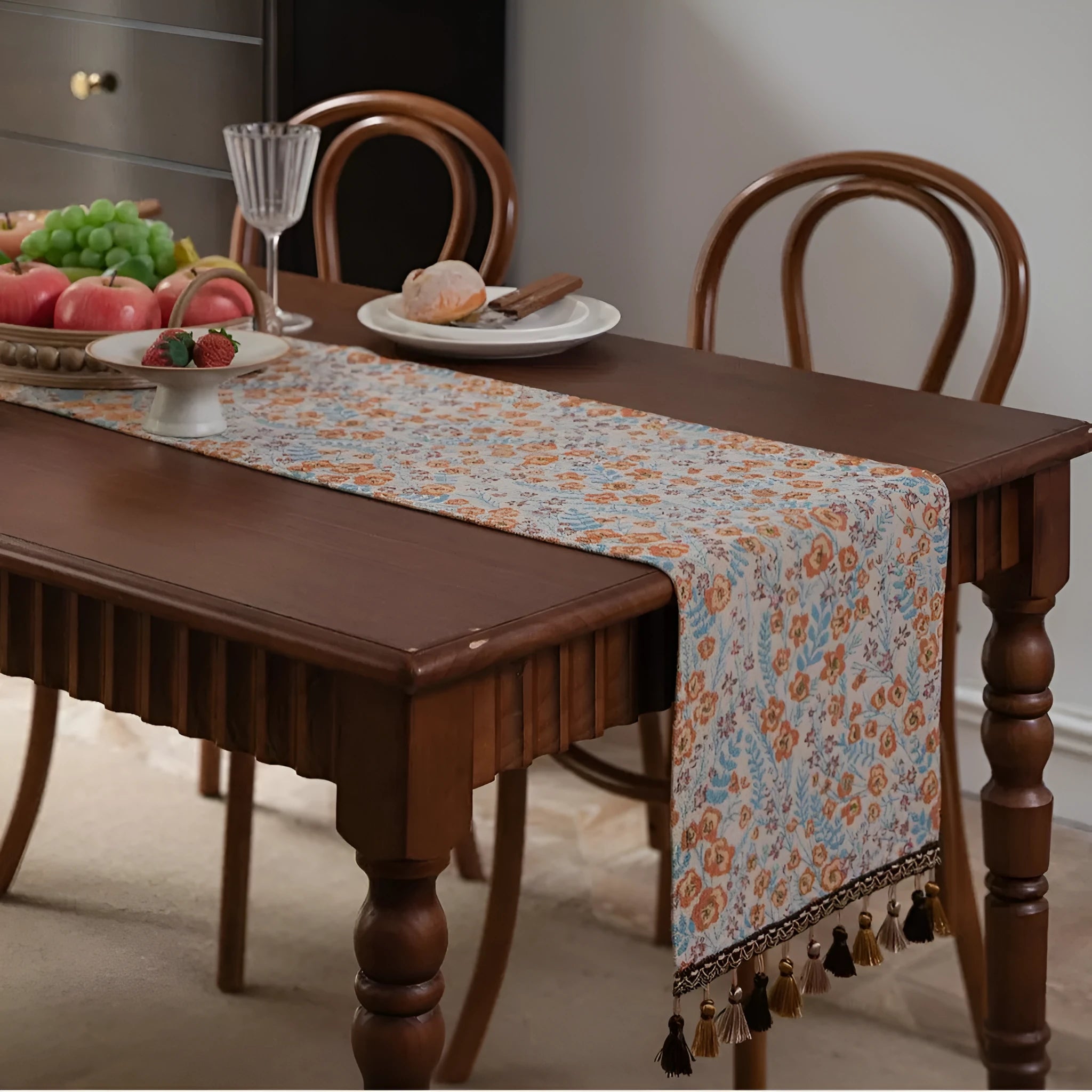 Long beige jacquard table runner with floral pattern and tassels on a wooden dining table.