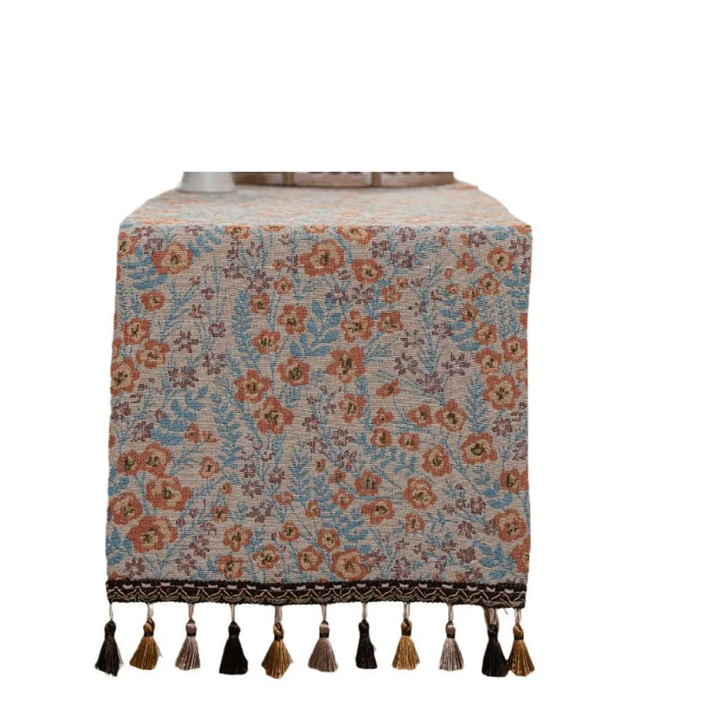 Long beige jacquard table runner with floral pattern and tassels on a wooden dining table.