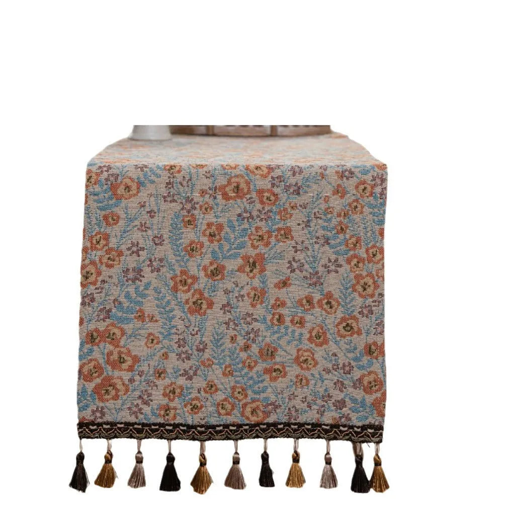 Long beige jacquard table runner with floral pattern and tassels on a wooden dining table.