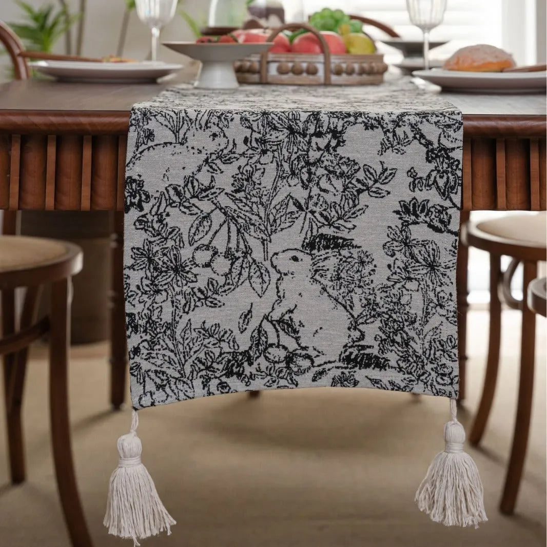 Long beige jacquard table runner with floral pattern and tassels on a wooden dining table.