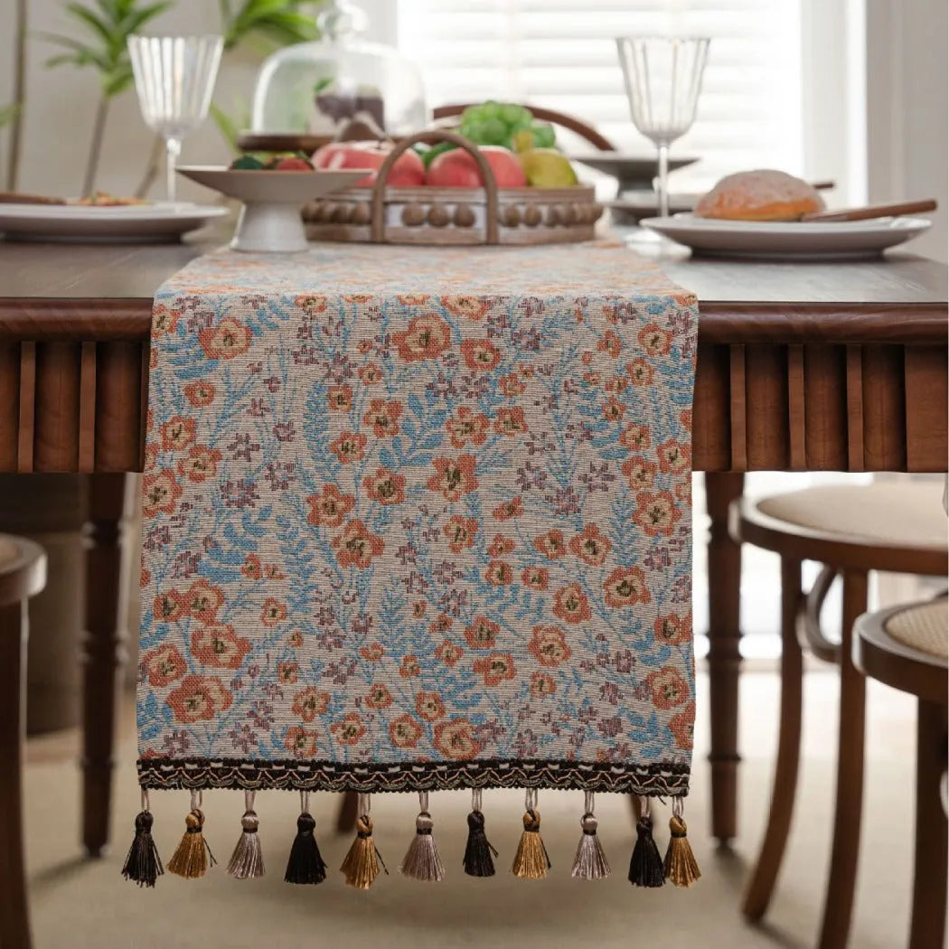 Long beige jacquard table runner with floral pattern and tassels on a wooden dining table.
